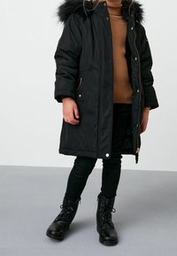 Parka noire à capuche avec bordure en fourrure, dotée de ferrures dorées, de poches zippées et d'une texture matelassée. Portée sur un pull côtelé marron et un pantalon noir.