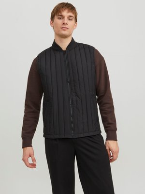 Gilet rembourré noir avec un col montant, fermeture éclair à l'avant et coutures verticales, porté sur une chemise à manches longues marron. Léger et texturé.
