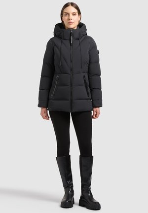 Femme portant une veste matelassée noire à capuche avec poches zippées, pantalon noir et bottes noires montant jusqu'aux genoux, debout sur un fond uni.