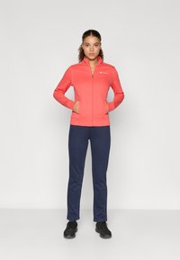 Jachetă coral cu fermoar, guler înalt, buzunare laterale și logo alb; asociată cu pantaloni bleumarin și încălțăminte sport neagră. Textură de țesătură netedă.