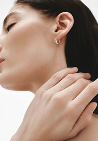 Aros de plata en forma de aro llevados en la oreja, con un diseño suave y redondeado. Tono de piel natural y mano delicada visibles en la imagen.