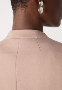 Light beige blazer with a structured collar, featuring a vertical seam and a small metal accent near the neck. Smooth texture.