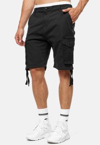 Shorts cargo noirs en coton, avec de multiples poches, des sangles réglables sur les côtés et une fermeture à bouton. Portés avec des baskets blanches et des chaussettes.