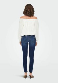 Balta off-the-shoulder blūze ar garām piedurknēm, apvienota ar tumši ziliem šauriem džinsiem, kuriem ir aizmugurējās kabatas un neliela stiepšanās.