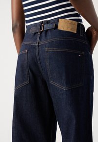 Osoba w ciemnoniebieskich dżinsach z tylnymi kieszeniami oraz tan skórzaną metką z napisem „Tommy Jeans”, w połączeniu z niebieską bluzką w paski.