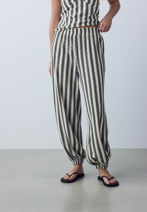 Woman wearing black and white vertical striped elastic-waist pants with elastic cuffs and black flip-flop sandals, standing on a gray floor.