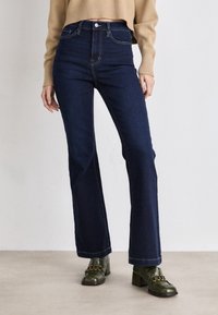 Visoko pasna, temno modra kavbojska flared jeans hlače s svetlim šivom in sprednjimi žepi, v kombinaciji z zelenimi čevlji na peto.