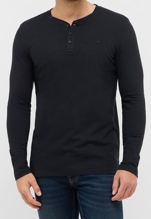 Homme portant un t-shirt henley noir ajusté à manches longues avec trois boutons et un jean bleu foncé sur un fond uni.
