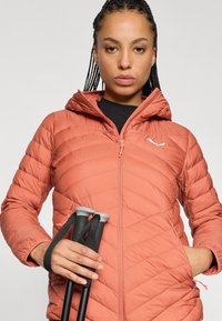 Casaco acolchoado em coral-laranja com fecho éclair na frente, capuz e padrão em ziguezague. Bastões de trekking pretos segurados numa mão.