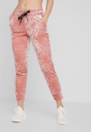 Femme portant un pantalon de jogging en velours écrasé rose clair avec un cordon noir et des baskets blanches, debout devant un fond clair uni.