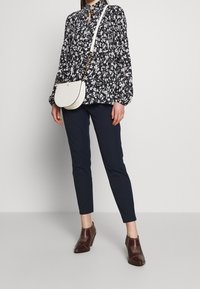 Blusa a maniche lunghe bianca e nera con motivi floreali e colletto con fiocco, abbinata a pantaloni aderenti blu navy e stivaletti marroni con tacco. Borsa a tracolla bianca.