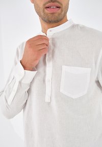 Uomo che aggiusta il colletto di una camicia bianca in lino con bottoni e taschino, viso inferiore e barba incolta in vista, sfondo bianco semplice.