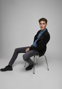 Schwarze Wildlederjacke über einem Denimhemd, graue Hose und schwarze Schuhe. Modell sitzt auf einem Metallstuhl vor einem grauen Hintergrund.