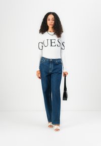 White long-sleeve shirt with "GUESS" logo, paired with blue wide-leg jeans. Model holds a small black handbag and wears light-colored open-toe shoes.
