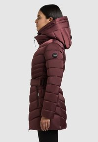 Vinasto rjava puhovka z kapuco, pasom v predelu pasu, žepi z zadrgo in rebrastimi manšetami. Izstopajoč vzorec z odejo.