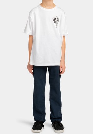 Camiseta blanca de manga corta con un pequeño diseño gráfico en el lado izquierdo del pecho, combinada con pantalones azul oscuro y zapatillas negras.