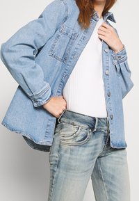 Light blue denim jacket with button closure and chest pocket, worn over a white ribbed top, paired with light blue jeans featuring faded details.