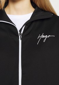 Chaqueta negra con cremallera y logo "Hugo" en blanco en el pecho. Hecha de una tela suave y texturizada.