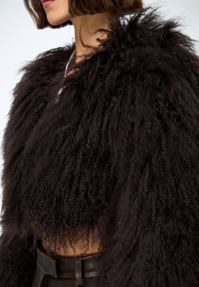 Zwarte, pluizige cropped jas gemaakt van getextureerd materiaal. Heeft lange, krullende vezels en een brede kraag. Draagt over een bruine, hooguitvallende ceintuur.