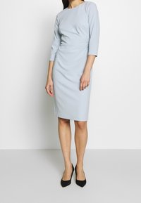 Ljusblå bodycon-klänning med ¾ ärmar, texturerad tyg, rund halsringning; stylar med svarta spetsiga klackar.