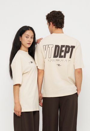 Twee mensen dragen beige T-shirts met de tekst "YT DEPT", één kijkt naar voren en de ander toont het ontwerp aan de achterkant. Beiden dragen donkere broeken.