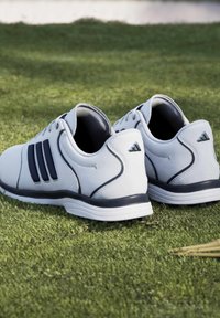 Ζευγάρι λευκά και μαύρα παπούτσια γκολφ Adidas τοποθετημένα σε πράσινο γρασίδι κοντά σε tee γκολφ.