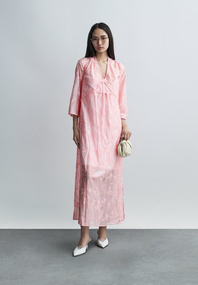 Μακριά ροζ φόρεμα με μανίκια 3/4, διακόσμηση από δαντέλα, V-λαιμόκοψη και λουλουδένια σχέδια. Το μοντέλο κρατά μια μικρή λευκή τσάντα και φορά λευκά mules με τακούνι.