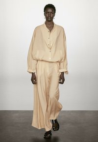 Ljusbeige oversized skjorta med knappar och vida byxor i textil. Tillsammans med svarta sandaler med remmar står modellen mot en neutral bakgrund.