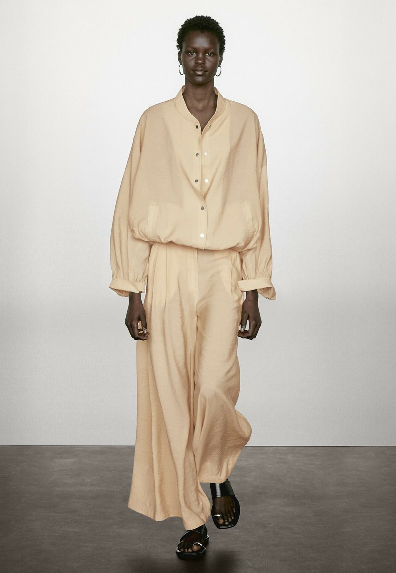 Ljusbeige oversized skjorta med knappar och vida byxor i textil. Tillsammans med svarta sandaler med remmar står modellen mot en neutral bakgrund.
