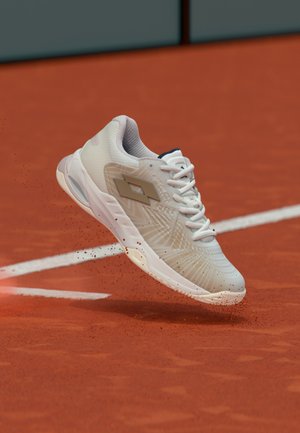 Balta sporta apava ar teksturētu augšdaļu, auklu detaļām un logotipu. Piekārta virs sarkanas māla virsmas, redzami nelieli putekļu daļiņas.