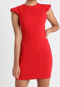 Czerwona sukienka bez rękawów z teksturowanej tkaniny, dopasowany krój i falbaniaste akcenty na ramionach. Gładkie wykończenie i długość powyżej kolan.