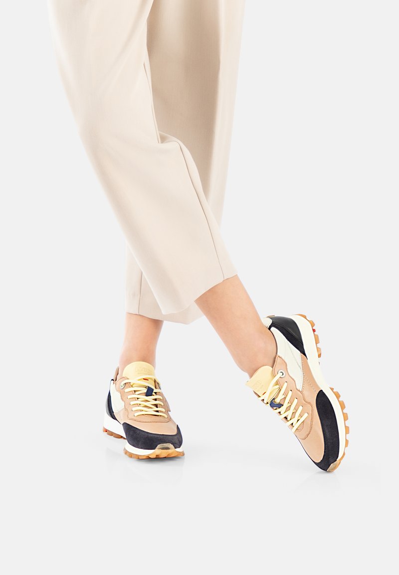 Sneakers with a mix of beige, black, and white materials. Features yellow laces and a textured sole for grip. Rounded, athletic design.
