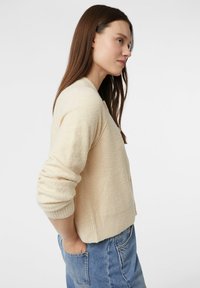 Jasnobeżowy, teksturowany sweter z okrągłym dekoltem i długimi rękawami. Połączony z niebieskimi dżinsami; luźny krój z delikatnymi prążkowaniami.