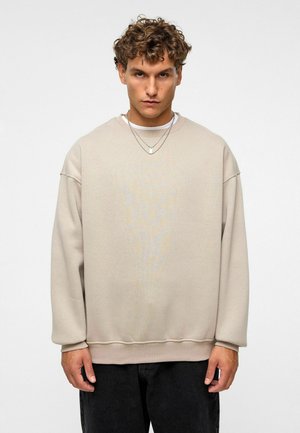 Beige oversized sweatshirt met een geprofileerde afwerking, verlaagde schouders en geribbelde manchetten, gedragen over een witte crew-neck shirt.