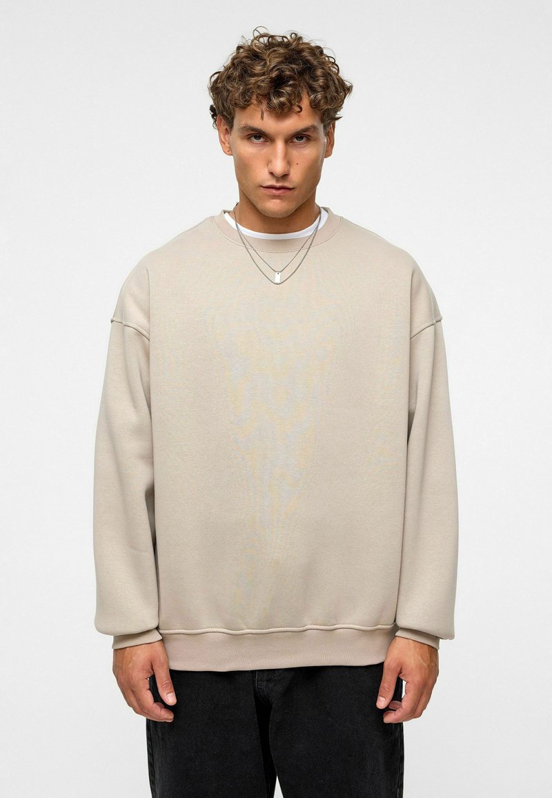Beige oversized sweatshirt met een geprofileerde afwerking, verlaagde schouders en geribbelde manchetten, gedragen over een witte crew-neck shirt.