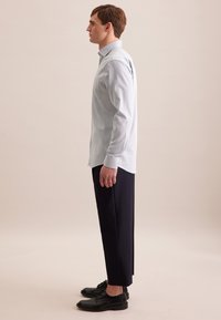Camicia azzurra chiara a maniche lunghe con taschino, abbinata a pantaloni scuri e sartoriali. Scarpe nere. Profilo laterale su uno sfondo neutro.