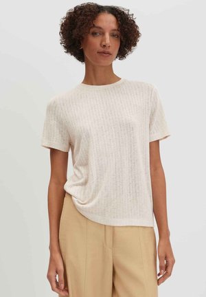 Donna con capelli corti e ricci che indossa una maglietta beige chiaro a maniche corte in maglia e pantaloni beige a vita alta, in piedi davanti a uno sfondo semplice.