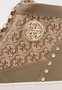 Gros plan d'une basket beige avec tissu monogramme beige, logo métallique doré, clous dorés et détails en cuir cousu beige.