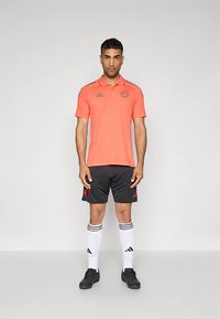 Tričko s krátkym rukávom v jasnej oranžovej farbe s golierom, v kombinácii s čiernymi kraťasmi a bielymi ponožkami s tromi čiernymi pruhmi.