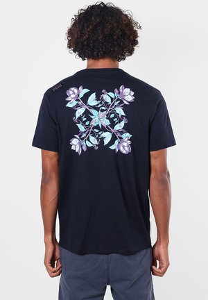 Homme portant un t-shirt noir avec un grand motif floral violet et bleu dans le dos, debout sur un fond clair uni.