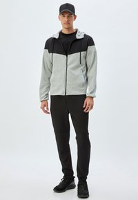 Grauer und schwarzer Zip-Hoodie mit Kapuze und Kordelzug, graue Jogginghose und schwarze Sportschuhe; glattes Material und taillierte Passform.