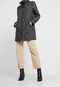 Cappotto in tweed bianco e nero con colletto alto, cerniera frontale completa, due tasche laterali e una vestibilità rilassata, abbinato a pantaloni cropped beige e stivali neri.