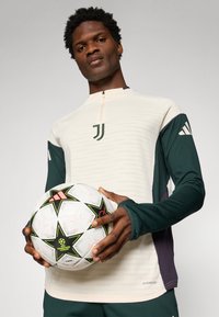 White long-sleeve top with green accents and zip neck, featuring Juventus logo. Player holds a white soccer ball with black and green star pattern.