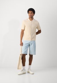 Beige knit polo shirt with short sleeves and buttons, light blue denim shorts, white sneakers, and a beige tote bag. Minimalistic design.