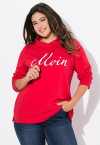 Felpa rossa con una grafica bianca "Moin", caratterizzata da un collo a cowl e maniche lunghe. Indossata con jeans blu scuro, che mostrano un look casual.