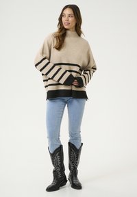 Beige és fekete csíkos, oversized pulóver magas nyakkal és hosszú ujjakkal, világoskék farmernadrággal és fekete cowboy csizmával párosítva.