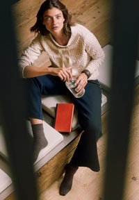 Kvinna sittande på en stoppad träbänk, klädd i en krämfärgad tröja och svarta byxor, håller ett glas vatten med en stängd röd bok intill.