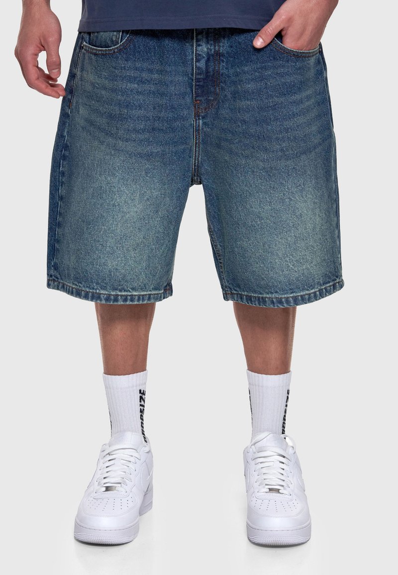 Homme portant un short en jean bleu, des chaussettes blanches montantes avec un texte noir et des baskets blanches à lacets, debout les mains près des poches.