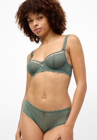Conjunto de lingerie verde em renda, apresentando um sutiã transparente com bordas onduladas e um detalhe de laço decorativo, combinado com uma cueca de renda correspondente.