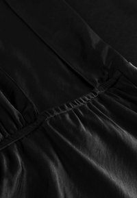 Čierny odev s hladkou, lesklou textúrou a nariasením v oblasti pásu. Látka je ľahká a jemne sa láme.